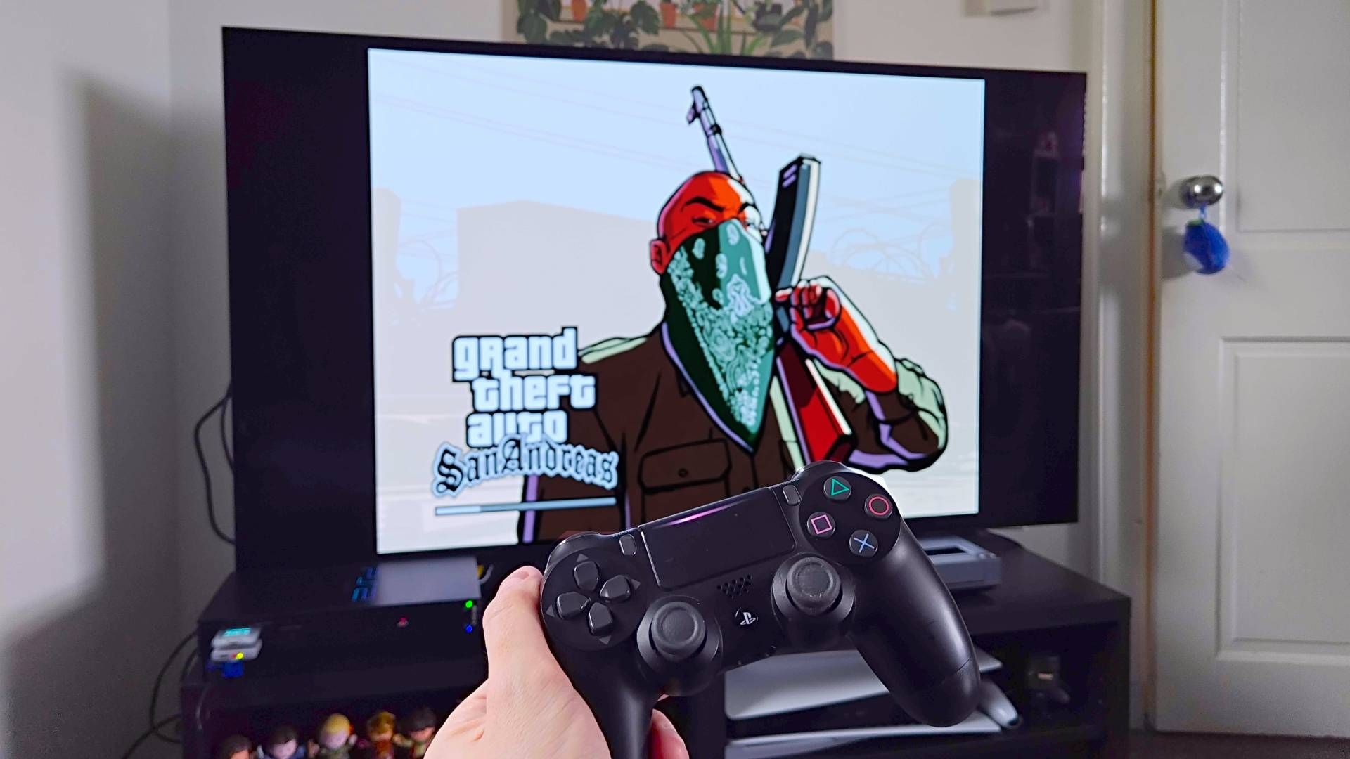 Hand holding DualShock 4 controller in front of PS2 connected to an LG OLED TV with GTA San Andreas load screen.